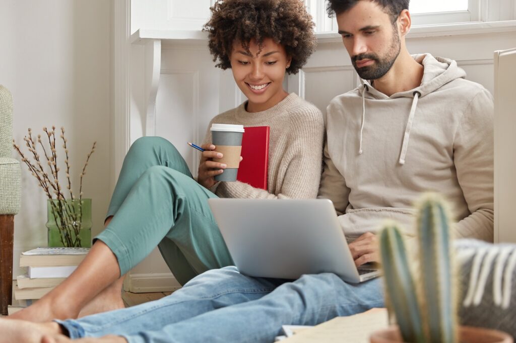 Image of family couple choose items for home in internet web store, make shopping online together, look at screen of laptop, drink espresso, sit on floor together. Technology and leisure concept